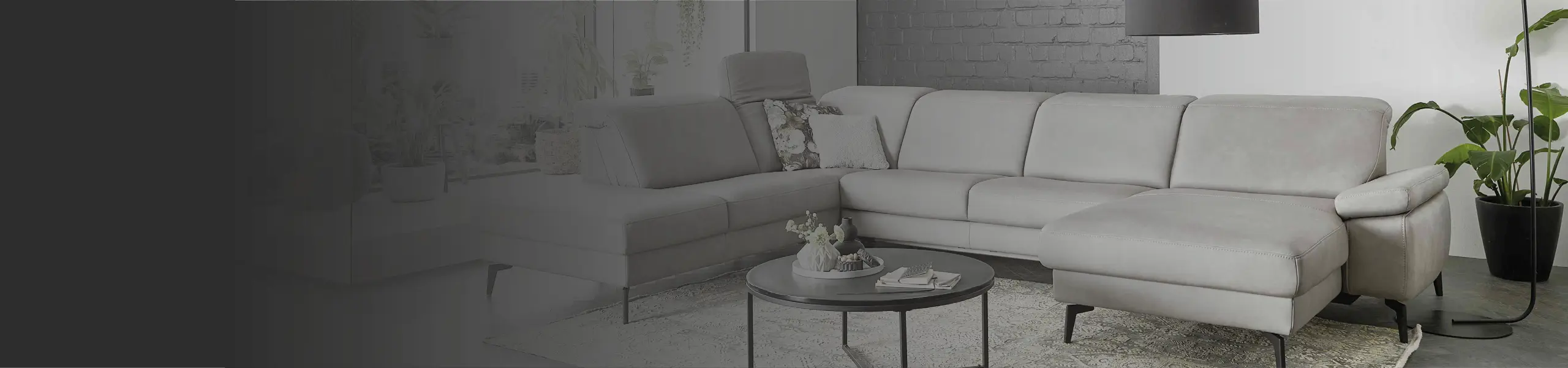 Stilvolles hellgraues Ecksofa mit verstellbaren Kopfstützen, gemütlichen Dekokissen und schwarzem Couchtisch im modernen Wohnzimmer mit Backsteinwand und Pflanzen. Perfekt für Wohnen mit Komfort und Design.